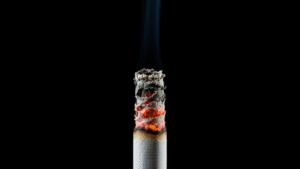 A close-up of a lit cigarette tip burning against a black background, releasing smoke and harmful chemicals.